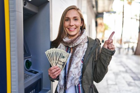 Photo for Young blonde woman holding dollars banknotes from atm machine smiling happy pointing with hand and finger to the side - Royalty Free Image