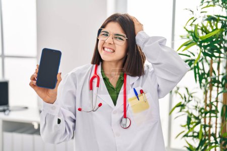 Téléchargez les photos : Jeune médecin hispanique femme montrant écran smartphone stressé et frustré avec la main sur la tête, visage surpris et en colère - en image libre de droit