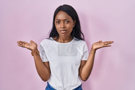 Foto de Mujer joven africana con camiseta blanca casual expresión despistada y confusa con los brazos y las manos levantadas. concepto de duda. - Imagen libre de derechos