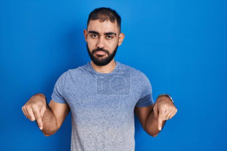 Photo for Middle east man with beard standing over blue background pointing down looking sad and upset, indicating direction with fingers, unhappy and depressed. - Royalty Free Image