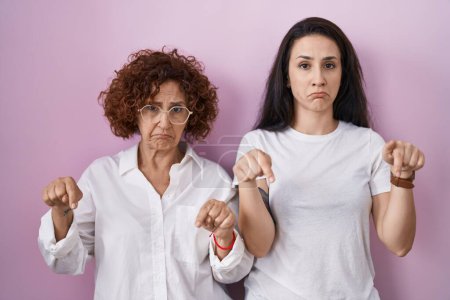 Foto de Madre e hija hispanas llevando camiseta blanca casual sobre fondo rosa apuntando hacia abajo buscando triste y molesto, indicando la dirección con los dedos, infeliz y deprimido. - Imagen libre de derechos