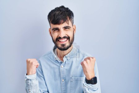 Téléchargez les photos : Jeune homme hispanique avec la barbe debout sur fond bleu très heureux et excité de faire geste gagnant avec les bras levés, souriant et criant pour le succès. concept de célébration. - en image libre de droit