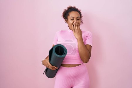 Foto de Mujer hispana joven con el pelo rizado sosteniendo estera de yoga sobre fondo rosa aburrido bostezo cansado cubriendo la boca con la mano. inquietud y somnolencia. - Imagen libre de derechos