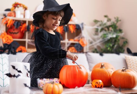 Foto de Adorable chica hispana teniendo fiesta de halloween poniendo dulce en cesta de calabaza en casa - Imagen libre de derechos