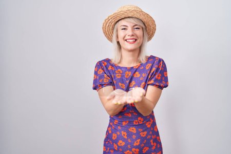 Foto de Mujer caucásica joven con vestido de flores y sombrero de verano sonriendo con las palmas de las manos juntas recibiendo o dando gesto. retención y protección - Imagen libre de derechos