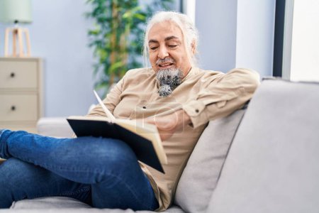 Foto de Hombre de pelo gris de mediana edad leyendo libro sentado en el sofá en casa - Imagen libre de derechos