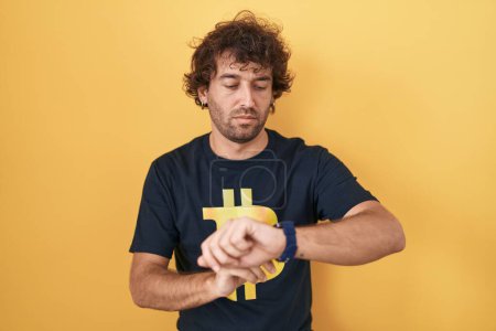 Foto de Joven hispano con camiseta bitcoin comprobando la hora en el reloj de pulsera, relajado y confiado - Imagen libre de derechos