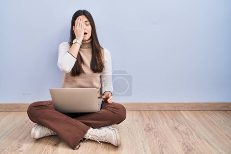 Téléchargez les photos : Jeune femme brune travaillant à l'aide d'un ordinateur portable assis sur le sol bâillant fatigué couvrant la moitié du visage, des yeux et de la bouche avec la main. mal au visage dans la douleur. - en image libre de droit