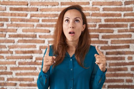 Photo for Brunette woman standing over bricks wall amazed and surprised looking up and pointing with fingers and raised arms. - Royalty Free Image