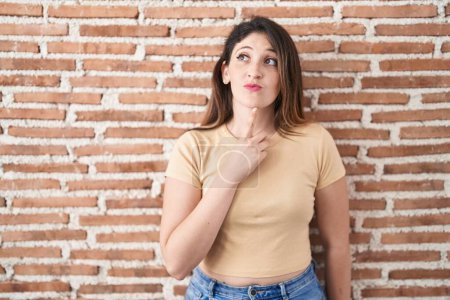 Photo for Young brunette woman standing over bricks wall thinking concentrated about doubt with finger on chin and looking up wondering - Royalty Free Image