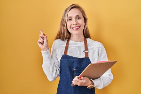Téléchargez les photos : Jeune femme blonde portant tablier de serveuse professionnelle tenant presse-papiers souriant heureux pointant avec la main et le doigt sur le côté - en image libre de droit