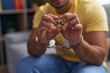 Young hispanic man criminal holding monero crypto currency wearing handcuffs at home