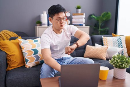 Téléchargez les photos : Jeune homme asiatique à l'aide d'un ordinateur portable à la maison assis sur le canapé en regardant le temps de la montre inquiet, peur de se tard - en image libre de droit