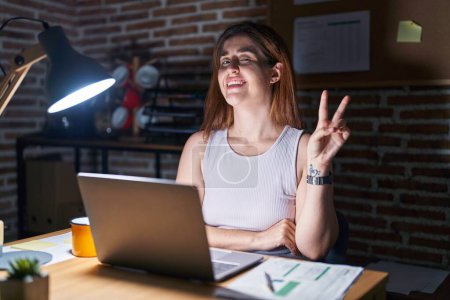 Téléchargez les photos : Femme brune travaillant au bureau la nuit souriant avec le visage heureux clin d'oeil à la caméra faisant signe de victoire. numéro deux. - en image libre de droit