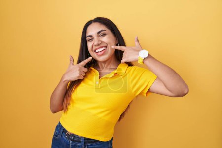 Téléchargez les photos : Jeune femme arabe debout sur fond jaune souriant gai montrant et pointant avec les doigts dents et la bouche. concept de santé dentaire. - en image libre de droit