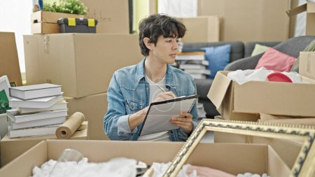Photo for Young hispanic man unpacking cardboard box writing on checklist at new home - Royalty Free Image