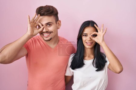 Foto de Joven pareja hispana de pie sobre fondo rosa haciendo buen gesto con la mano sonriente, los ojos mirando a través de los dedos con la cara feliz. - Imagen libre de derechos