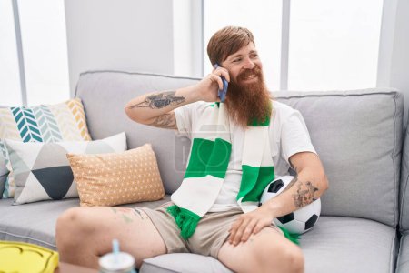 Photo for Young redhead man supporting soccer team talking on smartphone at home - Royalty Free Image