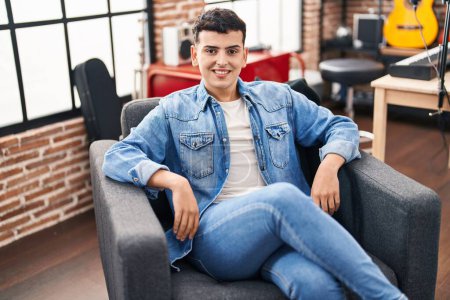 Téléchargez les photos : Jeune homme non binaire musicien souriant confiant assis sur la chaise au studio de musique - en image libre de droit