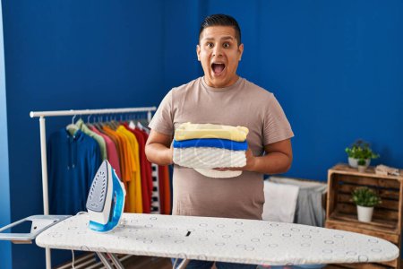 Photo for Hispanic young man holding folded laundry after ironing celebrating crazy and amazed for success with open eyes screaming excited. - Royalty Free Image