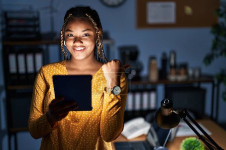 Photo for African american woman with braids working at the office at night with tablet pointing to the back behind with hand and thumbs up, smiling confident - Royalty Free Image