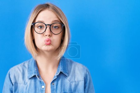 Téléchargez les photos : Young caucasian woman standing over blue background puffing cheeks with funny face. mouth inflated with air, crazy expression. - en image libre de droit