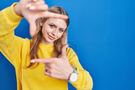 Téléchargez les photos : Jeune femme caucasienne debout sur fond bleu souriant faisant cadre avec les mains et les doigts avec le visage heureux. concept de créativité et photographie. - en image libre de droit