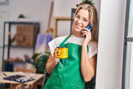 Téléchargez les photos : Jeune femme blonde parlant sur le smartphone buvant du café au studio d'art - en image libre de droit