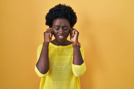 Foto de Mujer joven africana de pie sobre el estudio amarillo cubriendo las orejas con los dedos con expresión molesta por el ruido de la música alta. concepto de sordo. - Imagen libre de derechos