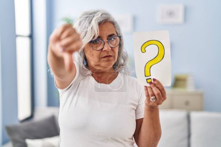 Foto de Mujer de mediana edad con pelo gris que sostiene signo de interrogación con la cara enojada, signo negativo que muestra aversión con los pulgares hacia abajo, concepto de rechazo - Imagen libre de derechos