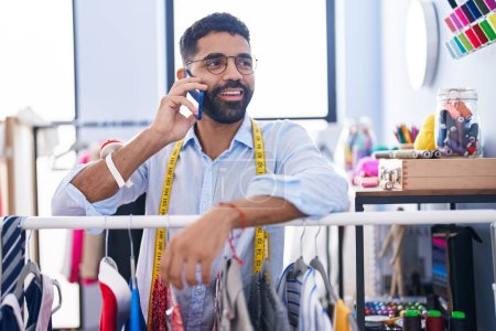 Téléchargez les photos : Jeune homme arabe tailleur parler sur smartphone appuyé sur le porte-vêtements au magasin de tailleur - en image libre de droit