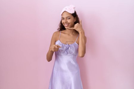 Photo for Young hispanic woman wearing sleep mask and nightgown smiling doing talking on the telephone gesture and pointing to you. call me. - Royalty Free Image