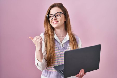 Téléchargez les photos : Jeune femme caucasienne travaillant à l'aide d'un ordinateur portable pointant vers le dos derrière avec la main et les pouces vers le haut, souriant confiant - en image libre de droit