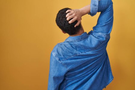 Photo for Hispanic man standing over yellow background backwards thinking about doubt with hand on head - Royalty Free Image