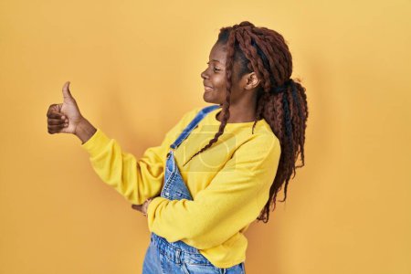 Foto de Mujer africana de pie sobre fondo amarillo mirando orgulloso, sonriendo haciendo pulgares hacia arriba gesto a un lado - Imagen libre de derechos