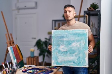 Photo for Young hispanic painter man holding canvas relaxed with serious expression on face. simple and natural looking at the camera. - Royalty Free Image