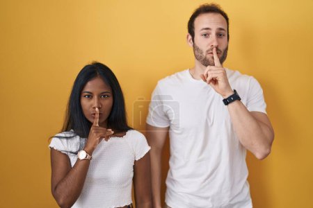 Téléchargez les photos : Couple Interracial debout sur fond jaune demandant à être calme avec le doigt sur les lèvres. silence et concept secret. - en image libre de droit