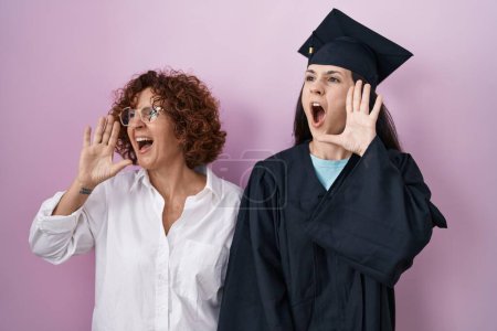 Foto de Madre e hija hispanas con gorra de graduación y bata de ceremonia gritando y gritando fuerte a un lado con la mano en la boca. concepto de comunicación. - Imagen libre de derechos