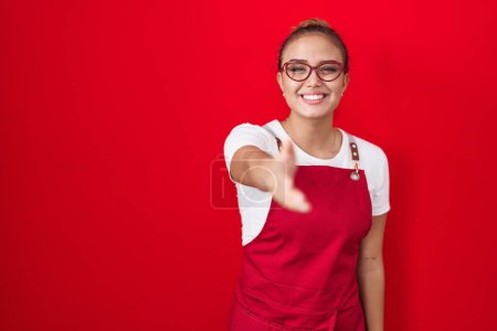 Photo for Young hispanic woman wearing waitress apron over red background smiling friendly offering handshake as greeting and welcoming. successful business. - Royalty Free Image