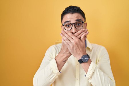 Photo for Hispanic young man wearing business clothes and glasses shocked covering mouth with hands for mistake. secret concept. - Royalty Free Image