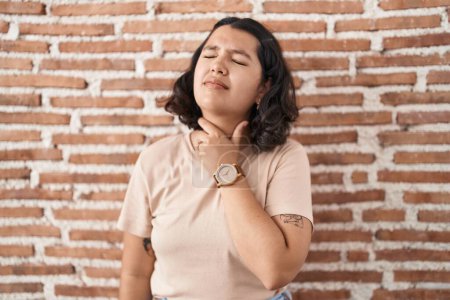Téléchargez les photos : Jeune femme hispanique debout sur un mur de briques touchant un cou douloureux, mal de gorge pour la grippe, la motte et l'infection - en image libre de droit