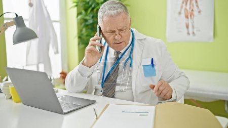 Photo for Middle age grey-haired man doctor talking on smartphone reading medical report at clinic - Royalty Free Image