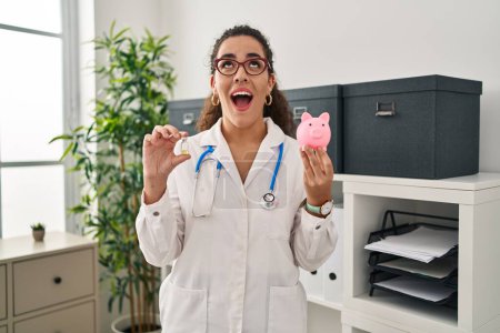 Photo for Young hispanic woman holding vaccine and piggy bank angry and mad screaming frustrated and furious, shouting with anger looking up. - Royalty Free Image