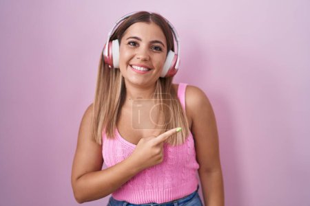 Foto de Mujer rubia joven escuchando música usando auriculares alegres con una sonrisa en la cara apuntando con la mano y el dedo hacia el lado con expresión feliz y natural - Imagen libre de derechos