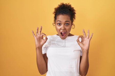 Photo for Young hispanic woman with curly hair standing over yellow background looking surprised and shocked doing ok approval symbol with fingers. crazy expression - Royalty Free Image