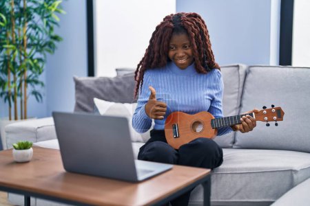 Téléchargez les photos : Femme africaine jouant ukulélé à la maison faire appel vidéo souriant heureux et positif, pouce levé faire excellent et signe d'approbation - en image libre de droit