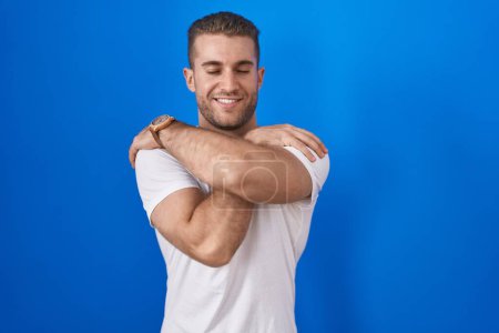 Photo for Young caucasian man standing over blue background hugging oneself happy and positive, smiling confident. self love and self care - Royalty Free Image