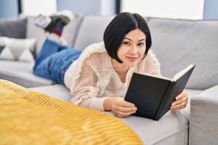Photo for Young chinese woman reading book lying on sofa at home - Royalty Free Image