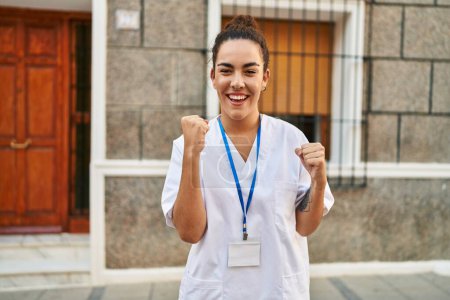 Téléchargez les photos : Jeune femme hispanique portant une carte d'identité de médecin célébrant surpris et étonnés pour le succès avec les bras levés et les yeux fermés - en image libre de droit