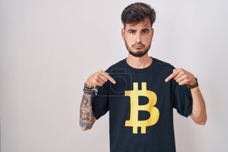 Foto de Joven hombre hispano con tatuajes con camiseta bitcoin apuntando hacia abajo buscando triste y molesto, indicando la dirección con los dedos, infeliz y deprimido. - Imagen libre de derechos